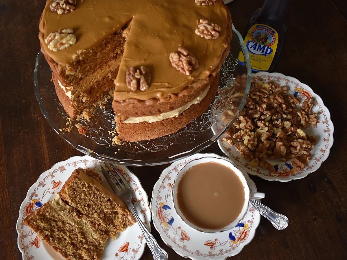 Coffee and Walnut cake on a cake stand with a slice of Coffee and Walnut Cake, cup of coffe, Camp Coffee bottle and a small plate of walnuts.